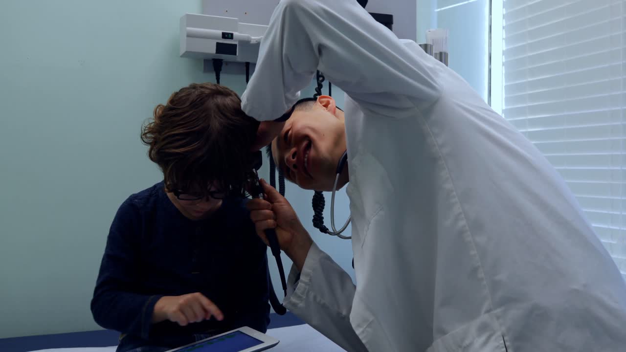 Side view of young asian male doctor examining a caucasian boy patient ear with tool in a clinic 4k
