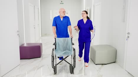 un médico hombre confiado con una barba gris en gafas en un uniforme azul rueda una silla para discapacitados y se comunica con una joven doctora morena que sostiene una tableta en sus manos a lo largo del corredor en una clínica moderna