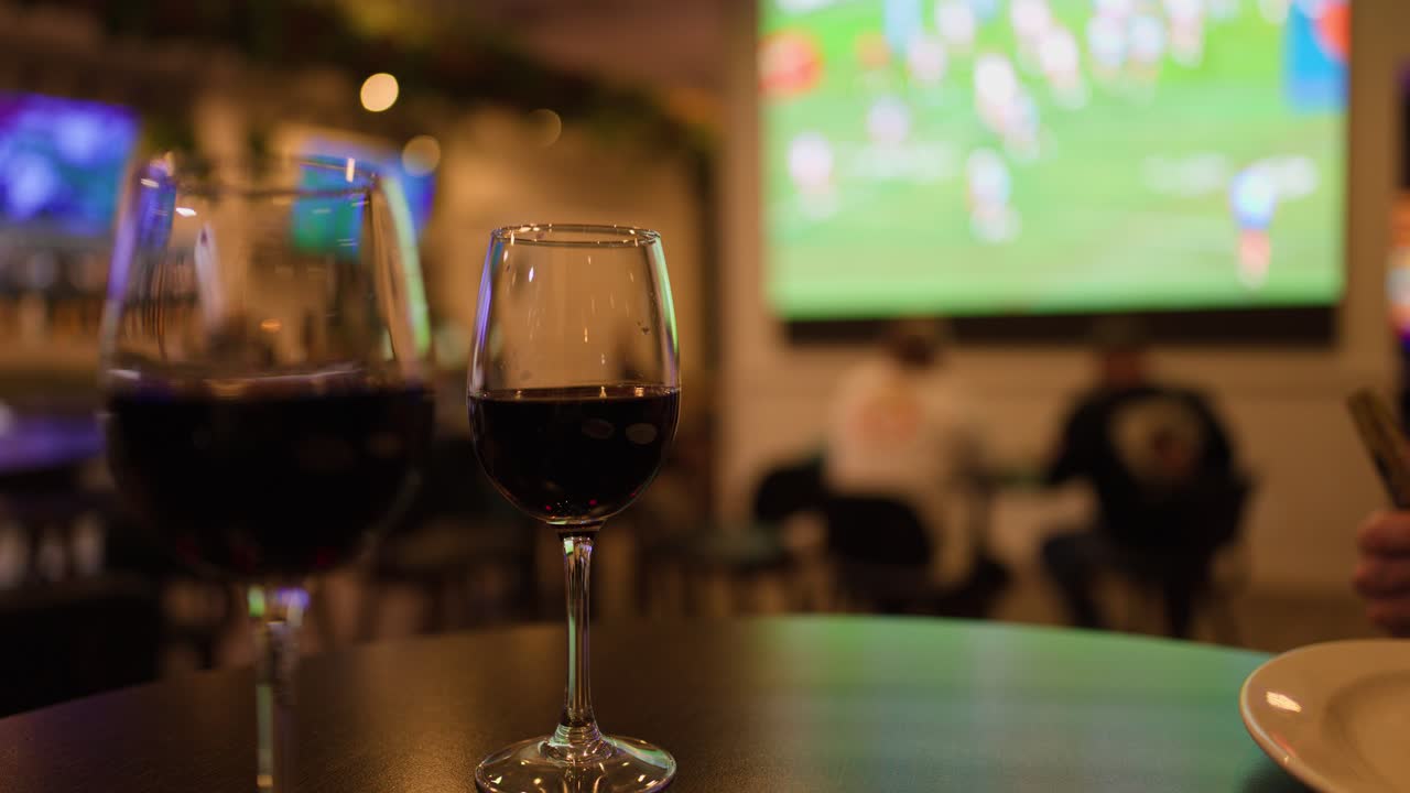 Hand places money beside wine glasses in dimly lit bar with televised sports game background