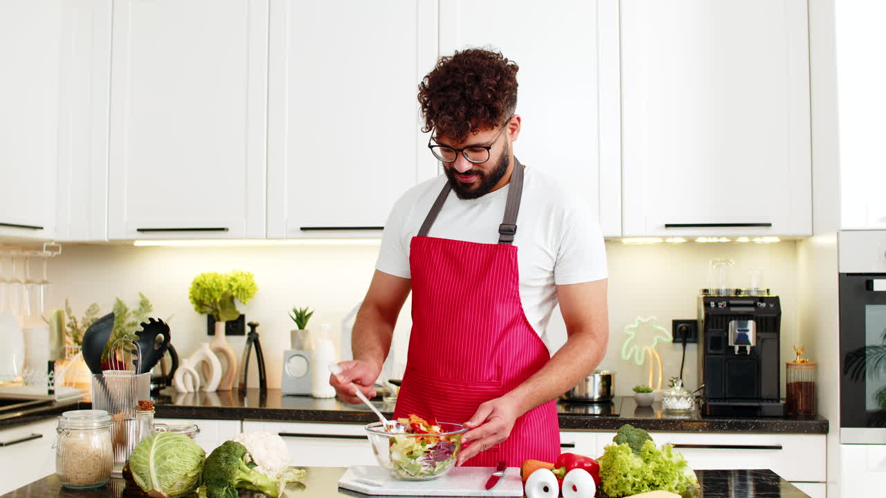 Giovane uomo arabo che mescola un'insalata fresca e salutare in cucina a casa, posizionando la ciotola sul tavolo per la preparazione del pasto.