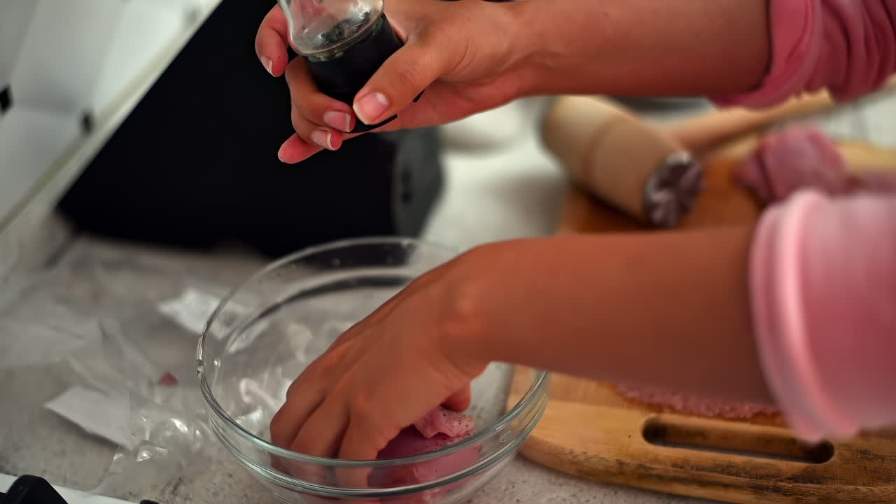 A hand sprinkles seasoning on raw chicken in a clear bowl, highlighting the careful preparation process in a tidy kitchen. The ingredients promise a flavorful dish