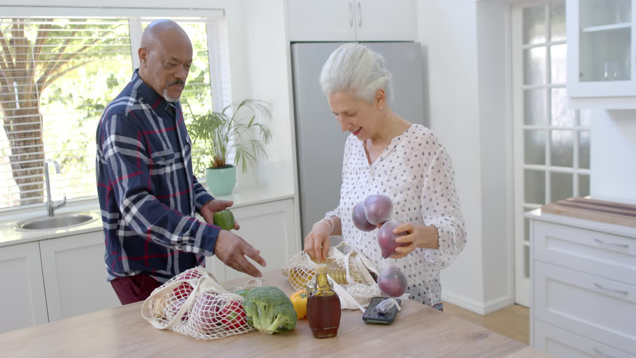 Casal birracial idoso feliz desempacotando sacolas de compras na cozinha, em câmera lenta