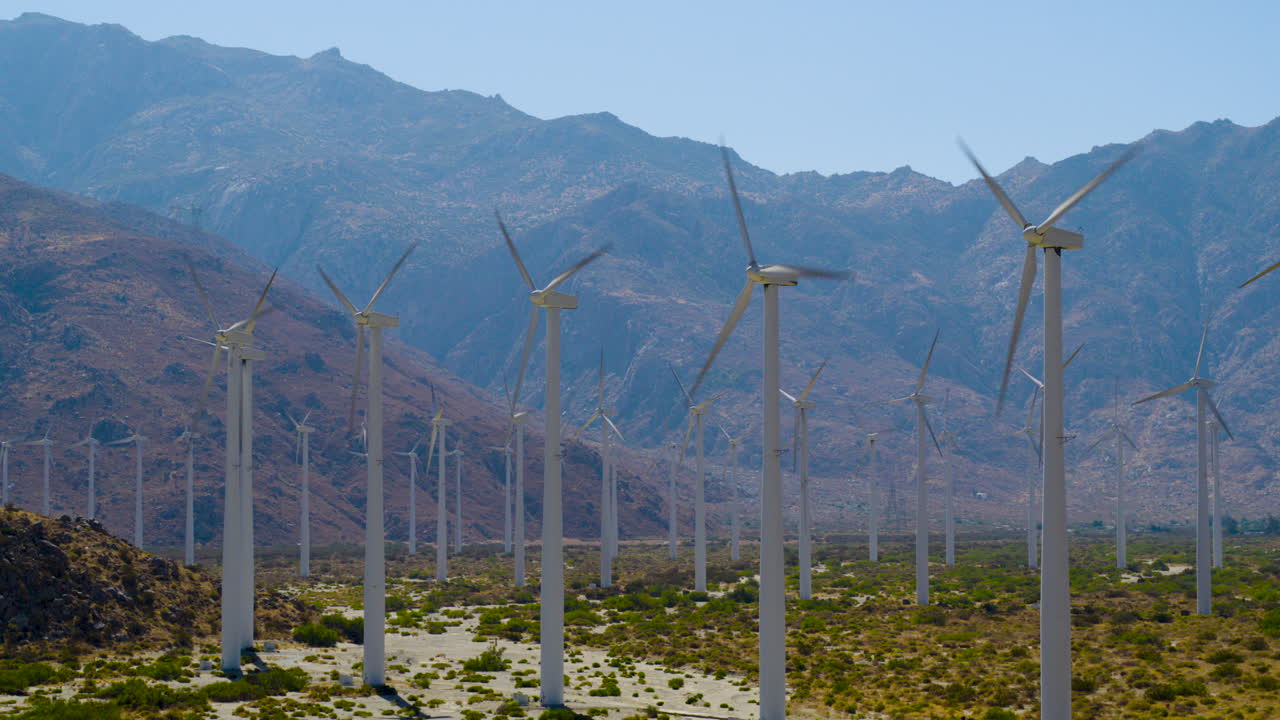 Palm Springs windmills spinning with the rugged San Jacinto Mountains rising behind them in California under a clear blue sky on a bright, sunny day. Aerial Shot spinning around them