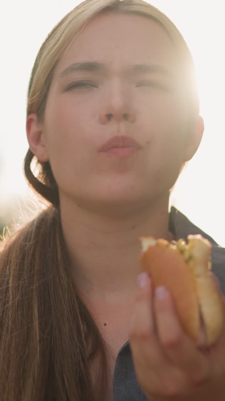 dama con camiseta gris disfruta de comer hamburguesa con una mirada de satisfacción, ojos cerrados, en un entorno al aire libre iluminado por el sol, el fondo borroso con los rayos del sol iluminando la escena