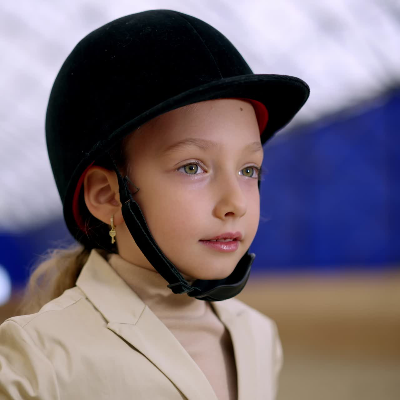 Adorable young kid wearing big black jockey helmet. Girl touching the cap smiling to the camera. Close up with gradual distancing. Blurred backdrop