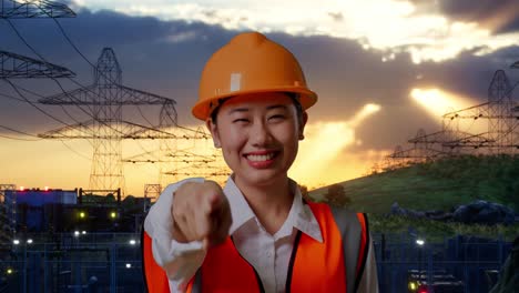 close up of asian female engineer with safety helmet smiling and touching her chest then pointing at you while standing near high voltage tower