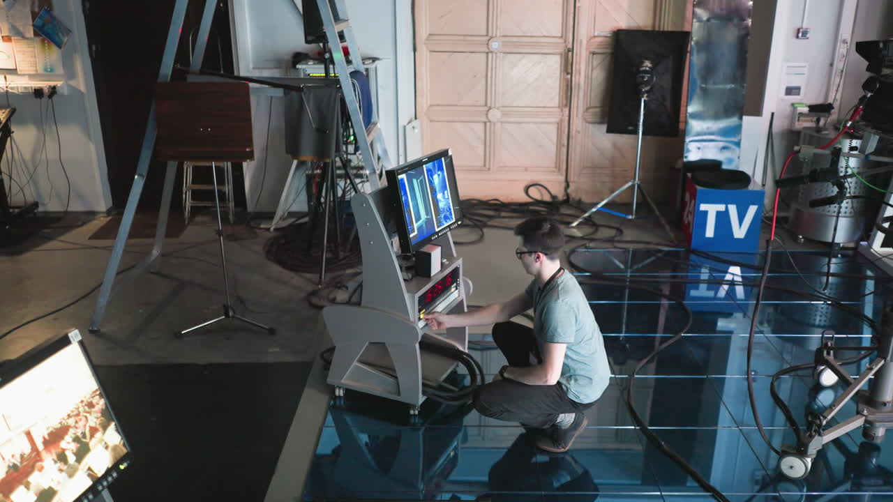 Man crouching and adjusting equipment in modern studio setting, timer visible on screen, filming gear in background, production action in process, tech-focused workspace