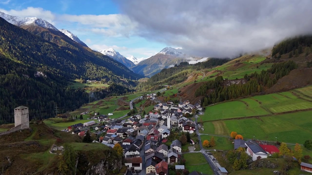 Volo lento di drone su un paese svizzero in autunno. Valle verde e montagne innevate sullo sfondo. Inquadratura ampia. Case e abitazioni in un piccolo villaggio svizzero. Strada e sentiero escursionistico sulla collina
