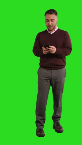 vertical video full length studio shot of businessman using mobile phone to message or browse online standing against green screen 2