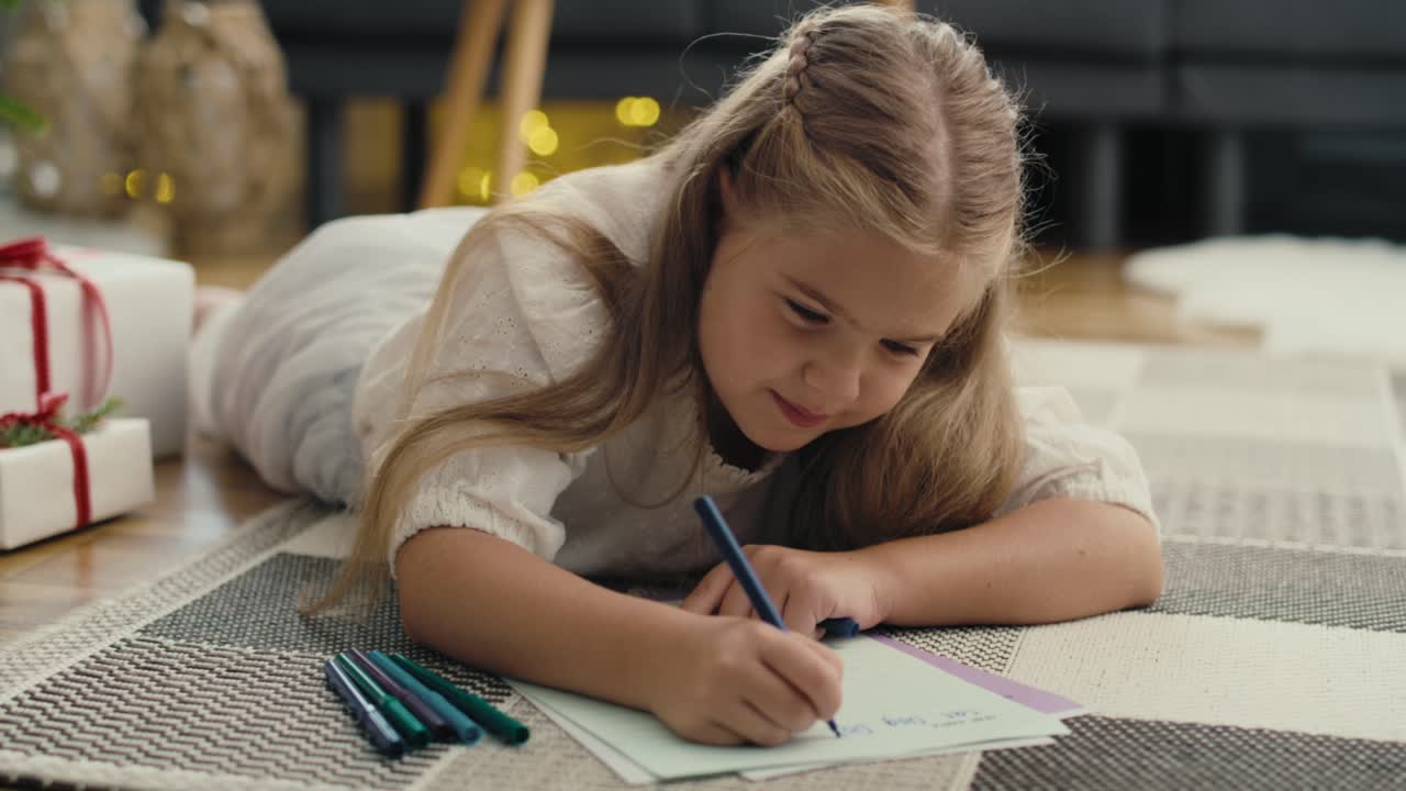 Close up of little caucasian girl lying on carpet next to Christmas tree and writing a letter to Santa Claus.