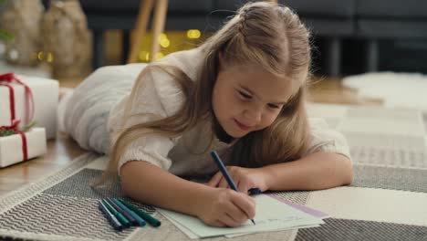 close up of little caucasian girl lying on carpet next to christmas tree and writing a letter to santa claus.