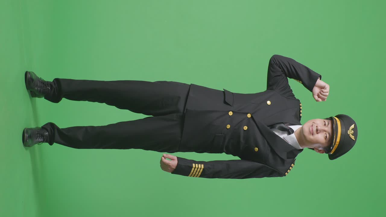 녹색 스크린 배경 스튜디오에 서 있는 동안 미소 짓고 팔다리를 구부리는 아시아 남성 조종사의 몸이 가득 차 있습니다.