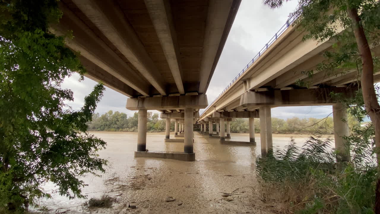 Bajo el puente, el río fluye lento y marrón, con pilares de hormigón que se desvanecen en la distancia mientras la naturaleza y la infraestructura se superponen en un tranquilo equilibrio.