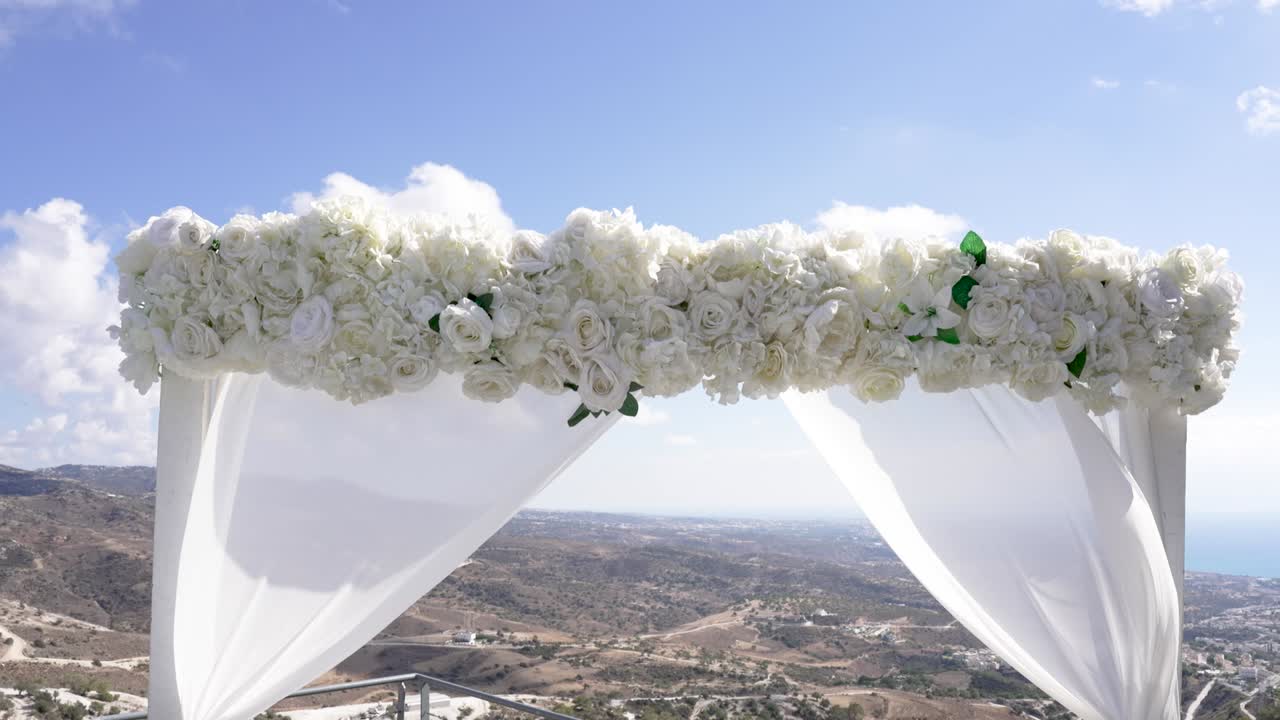 Un altare nuziale fatto di fiori bianchi si erge su una montagna. La vista mostra il mare e le colline. Il cielo è azzurro con qualche nuvola. Questa location è ideale per le cerimonie.