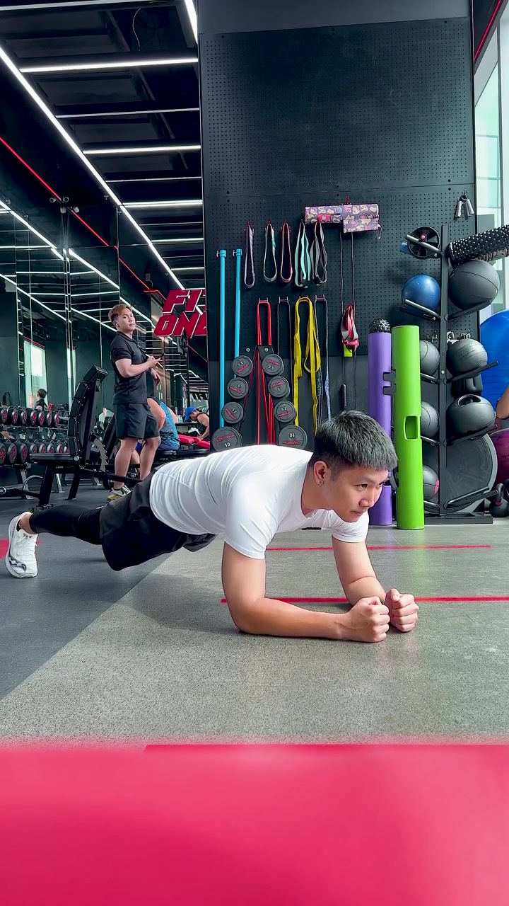 Joven realiza el ejercicio de plancha en una colchoneta en un gimnasio moderno, con iluminación brillante y ángulo de cámara estático.