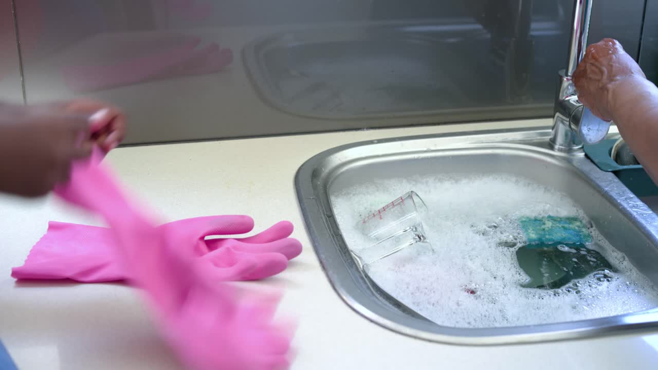 As adult hands are scrubbing at sink, child is pulling pink gloves on to help wash