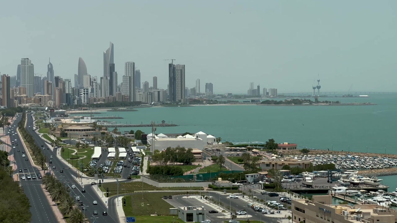 Vista ampla estática do horizonte da Cidade do Kuwait com marina na baía à beira-mar e estrada costeira durante o dia.