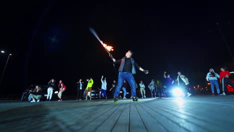 party outdoors with fire show and dances. young people watch the man shooting fire and hip-hopping near the race cars.