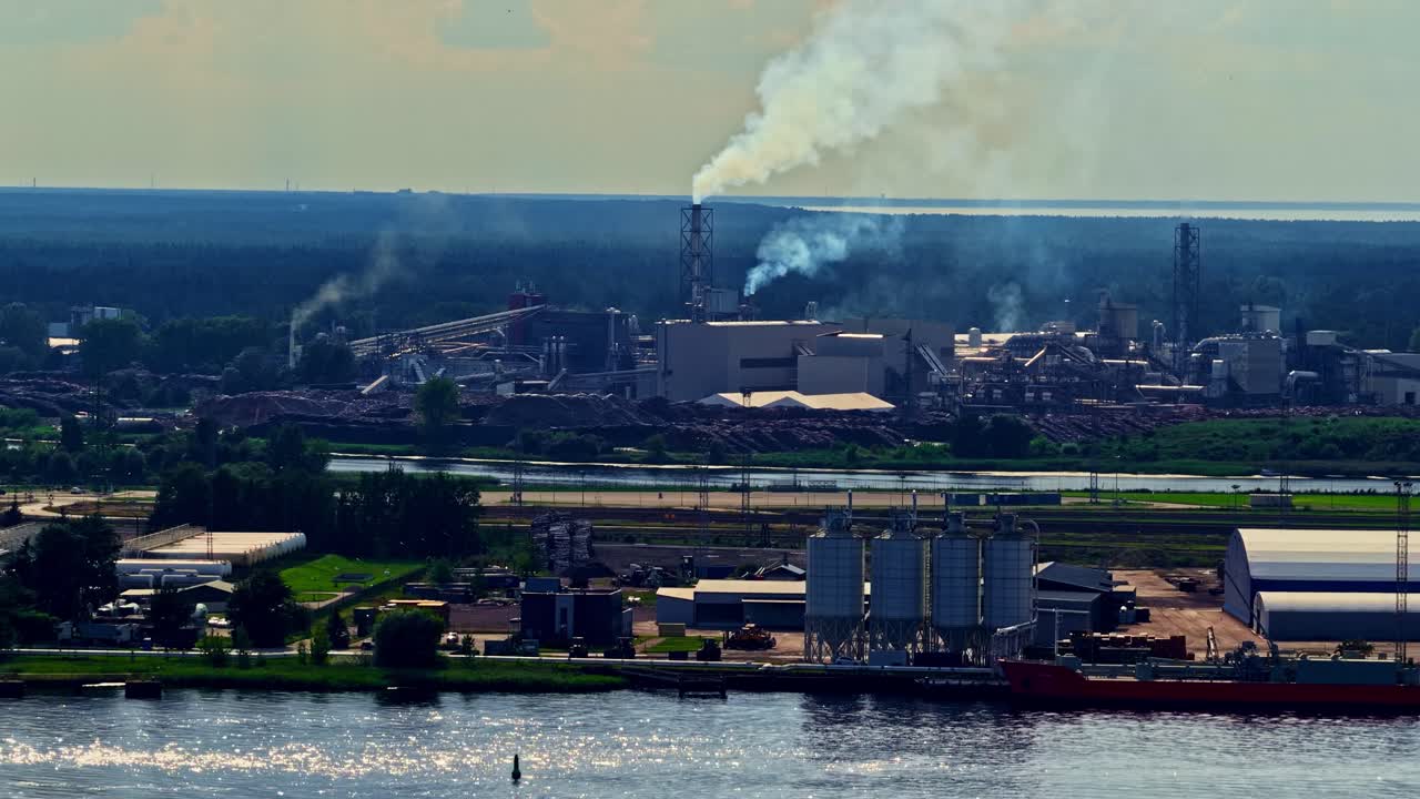 A wide aerial shot flies along the riverbank of the industrial Port of Riga, Latvia, showing a cargo ship, silos, and a large factory with smoke coming from its smokestacks