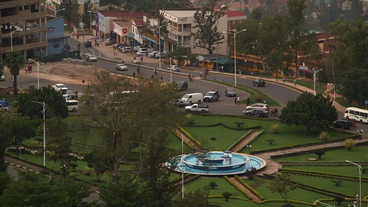 Vista dall'alto di un parco rotonda a Kigali, Ruanda, con fontana blu e giardini paesaggistici mentre il traffico cittadino scorre. Vita urbana, auto e pedoni