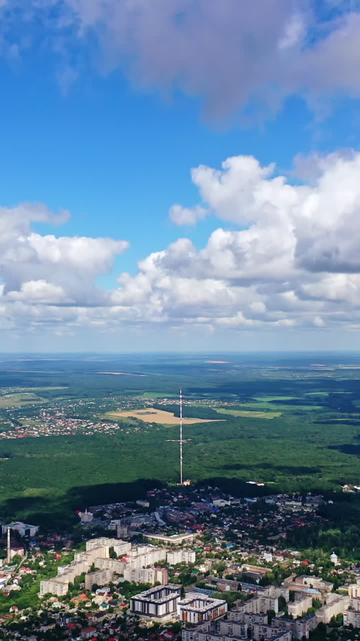 Panoramic view of a city among green nature. Cityscape under blue sky with white clouds. Beautiful scenery of a town near fields. Aerial view. Vertical video