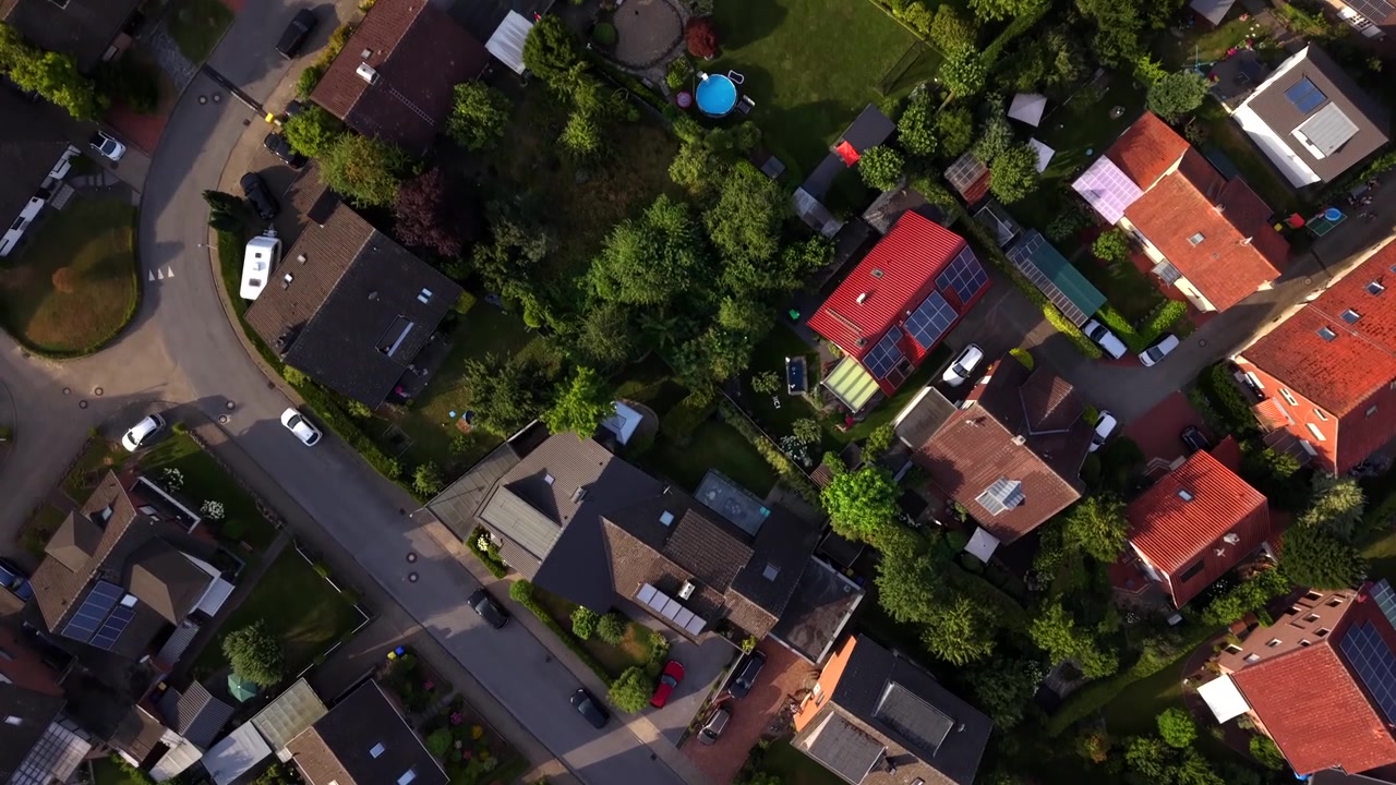 Quartiere residenziale tedesco con case unifamiliari dotate di pannelli solari e tetti in tegole rosse, auto parcheggiate lungo tranquille strade intersecanti illuminate dalla luce dorata della sera. Sorvolo aereo dall'alto con drone