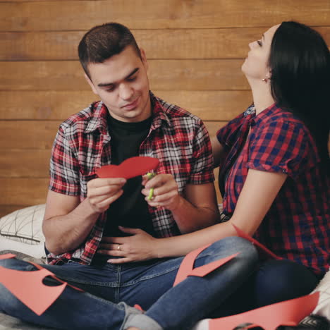 handsome man is cutting out heart from the red paper for his girlfriend over the wooden background. happy couple sitting on bed and the girl hugging and kissing her boyfriend.