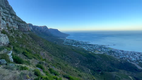 Amanecer-Caminata-Por-La-Mañana-Explorando-La-Montaña-De-La-Mesa-Ciudad-Del-Cabo-Sudáfrica-Pan-Izquierda-Vista-Del-Acantilado-De-La-Montaña-Y-El-Centro-Amarillo-Dorado-Rayos-De-Sol-Exuberante-Primavera-Verano-Hierba-Y-Flores-Azul-Profundo-Océano-Acuático