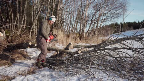 man using power saw to cut wood log outdoors in winter - wide shot