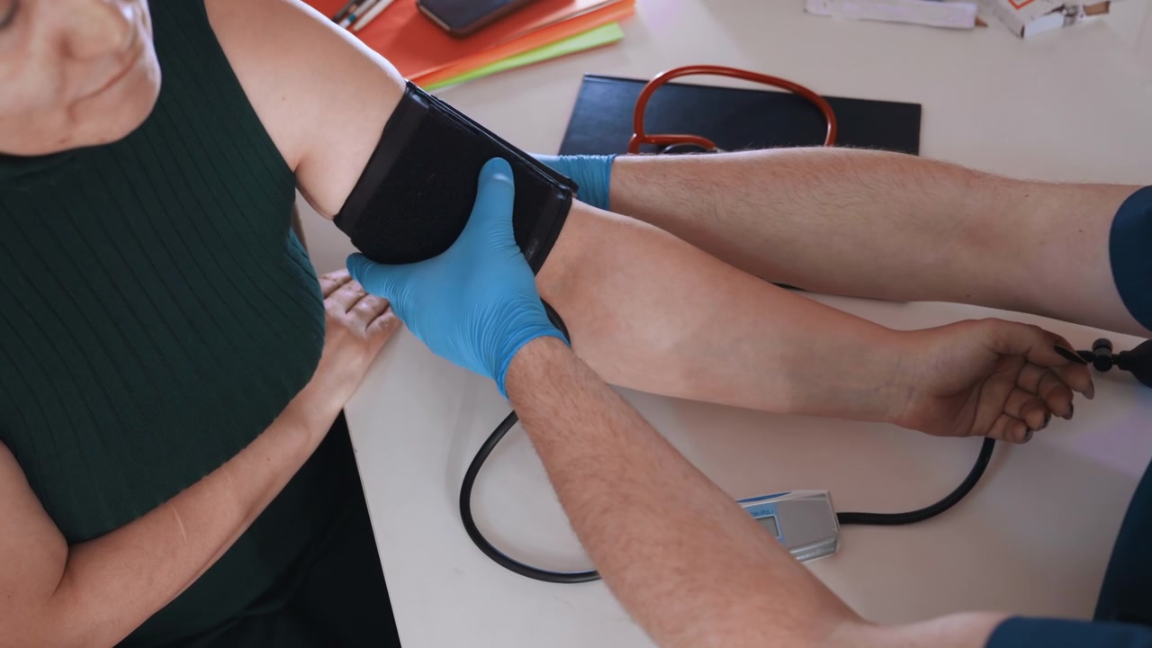 Monitoraggio del braccio e controllo del dispositivo che mostra bracciale, tubi e display digitale, il clinico maneggia lo strumento e registra la lettura, ambiente interno calmo con stetoscopio sul tavolo, verifica metodica.