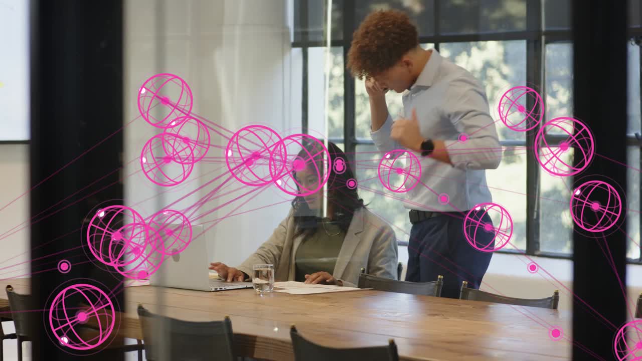 Standing man leaning pointing docs and guiding business woman reading laptop, pink overlay on glass