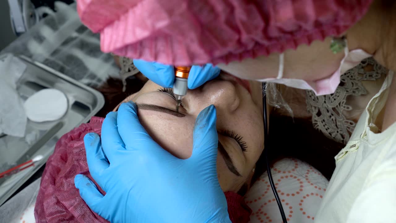 The girl lies on the couch in the beauty salon, and a professional beautician makes permanent makeup of her eyebrows. Microblading procedure. Close up. The view from the top. 4K, 25 fps.