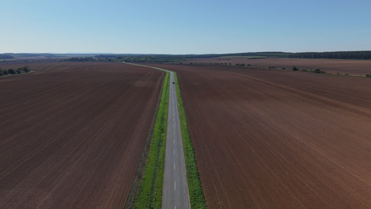 En flygvideo av en lång, rak landsväg som skär genom vidsträckta plöjda fält. Landskapet sträcker sig till horisonten med jordbruksmark på båda sidor, vilket visar upp lantlig enkelhet och öppna ytor.