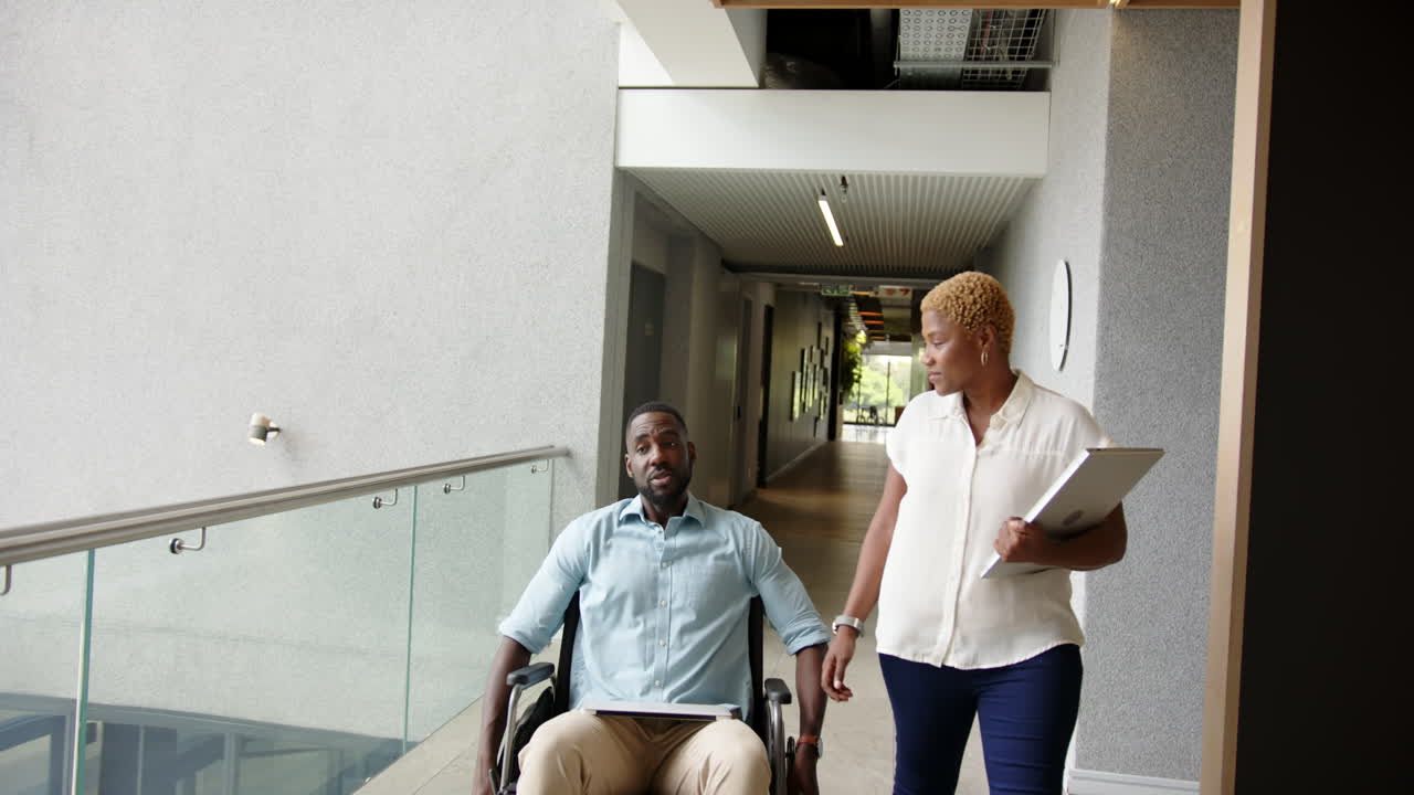 Discussing work, colleagues one using wheelchair with tablet and documents walking in office hallway