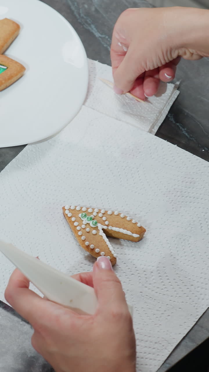 여성이 아이싱으로 쿠키를 장식하고 다른 쿠키가 트레이, 가위 및 장식 꽃에있는 동안 치아를 사용하여 녹색 구슬을 조정하는 위에서 아래로 볼 수 있습니다.