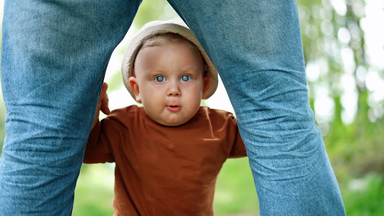 Cute baby boy in a cap stands holding by his dad's legs. Kid standing unsteadily learning to walk.