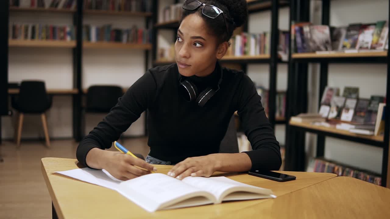 atractiva joven concentrada sentada junto al escritorio y haciendo algunas notas en su cuaderno para la universidad en la biblioteca moderna, preparándose para los exámenes