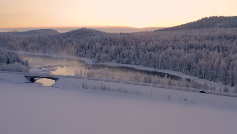 Luftaufnahme-Eines-Schnellen-Autos,-Das-über-Die-Eisseebrücke-Unter-Glühenden,-Verschneiten-Waldbergen-Bei-Sonnenaufgang-Fährt