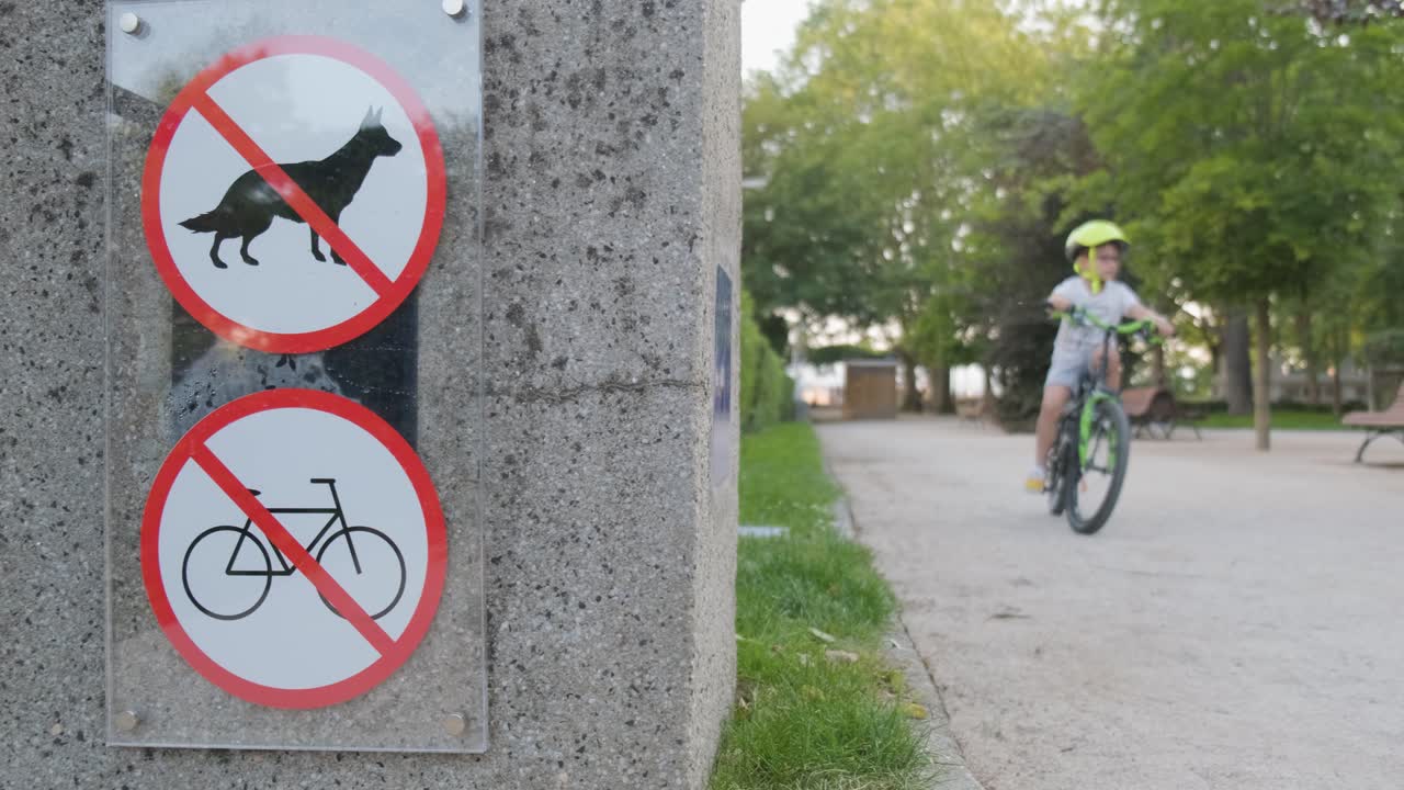 Young caucasian boy rides bike in park with no bicycles sign, dolly shot