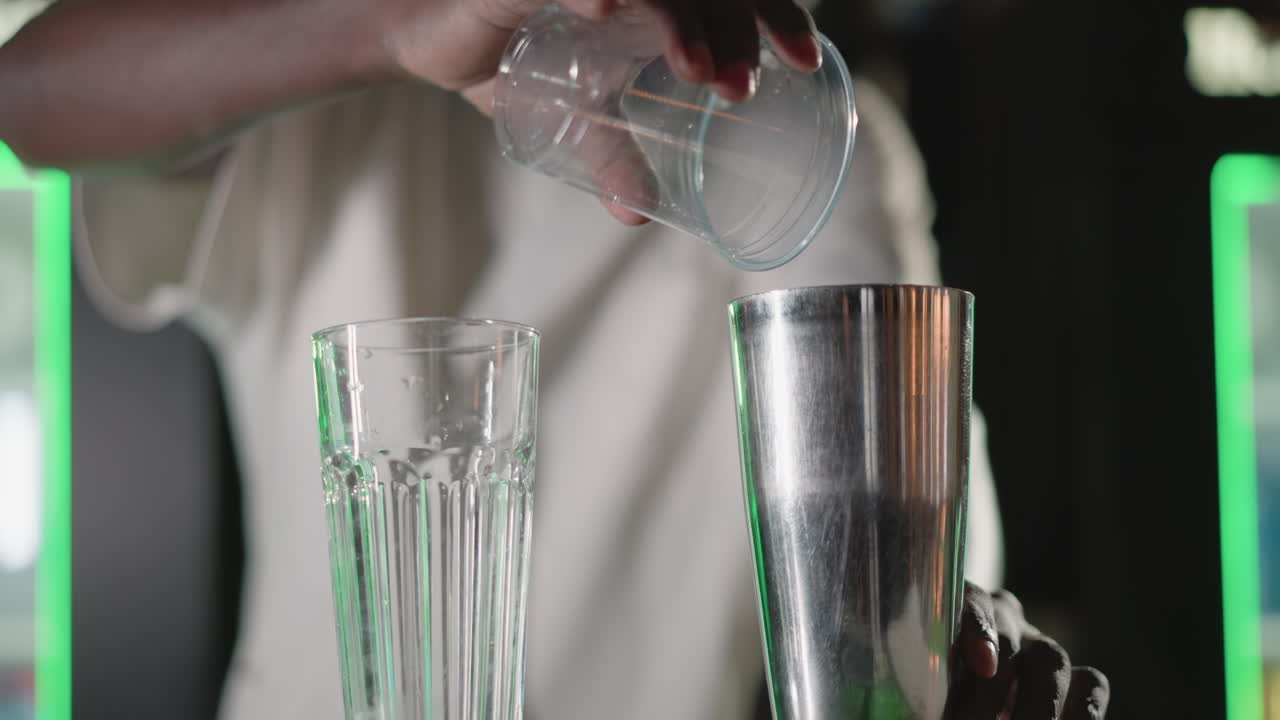 Bartender setting up glass and cocktail shaker on bar counter in modern bar with green lighting, preparing for drink mixing in vibrant nightlife atmosphere