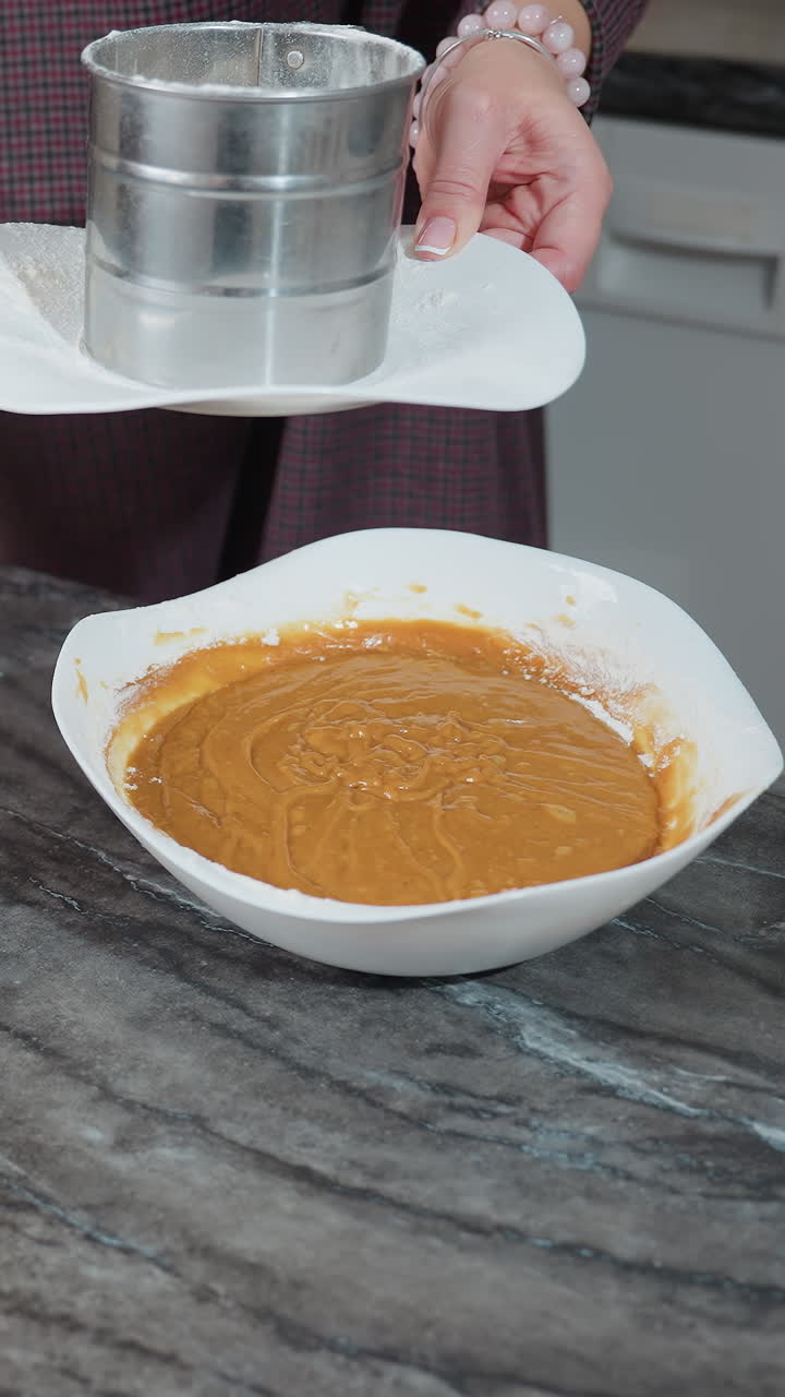 vista parcial de una mujer limpiando la encimera de la cocina con un paño mientras lleva un tamiz en el plato, un cuenco de masa se sienta en la superficie de mármol, destacando la preparación de alimentos y la limpieza en la cocina organizada