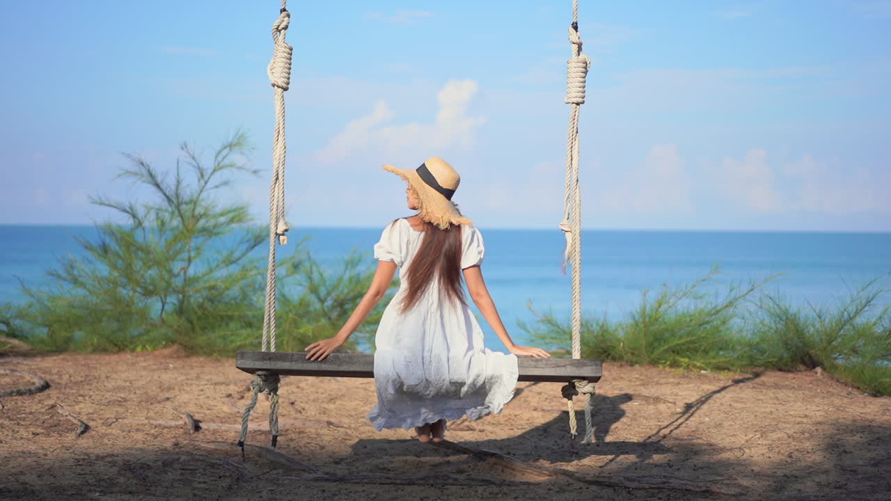 Young woman sitting and swinging on a wooden swing hanging on a tree at a tropical beach in Thailand enjoying the sea view, slow motion
