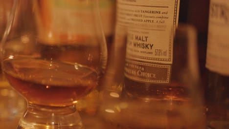 a hand gently swirls a whisky glass on a wooden bar, surrounded by bottles, under soft, warm ambient lighting with a shallow depth of field