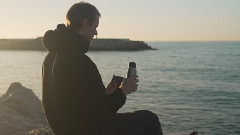 man-opens-thermos-pours-hot-drink-in-cup,-drinks-during-golden-hour