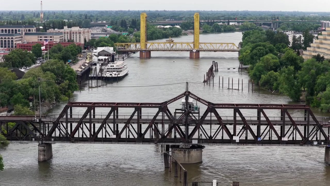 Fiume Sacramento con uno storico ponte a traliccio in acciaio e l'iconico Tower Bridge giallo che attraversano acque calme e marroni, con l'edificio a gradoni Ziggurat che si erge sopra le rive verdi del fiume e lo skyline urbano. Vista aerea.