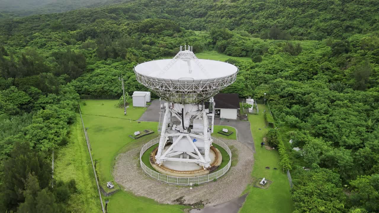 An aerial view captures a large radio telescope and an adjacent facility, situated in a clearing within a dense, lush green forest on an overcast day