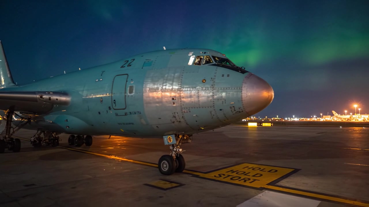 Sebuah pesawat menakjubkan menunggu keberangkatan di bawah tarian Aurora Borealis yang memukau di langit malam, di tengah latar bandara perkotaan.