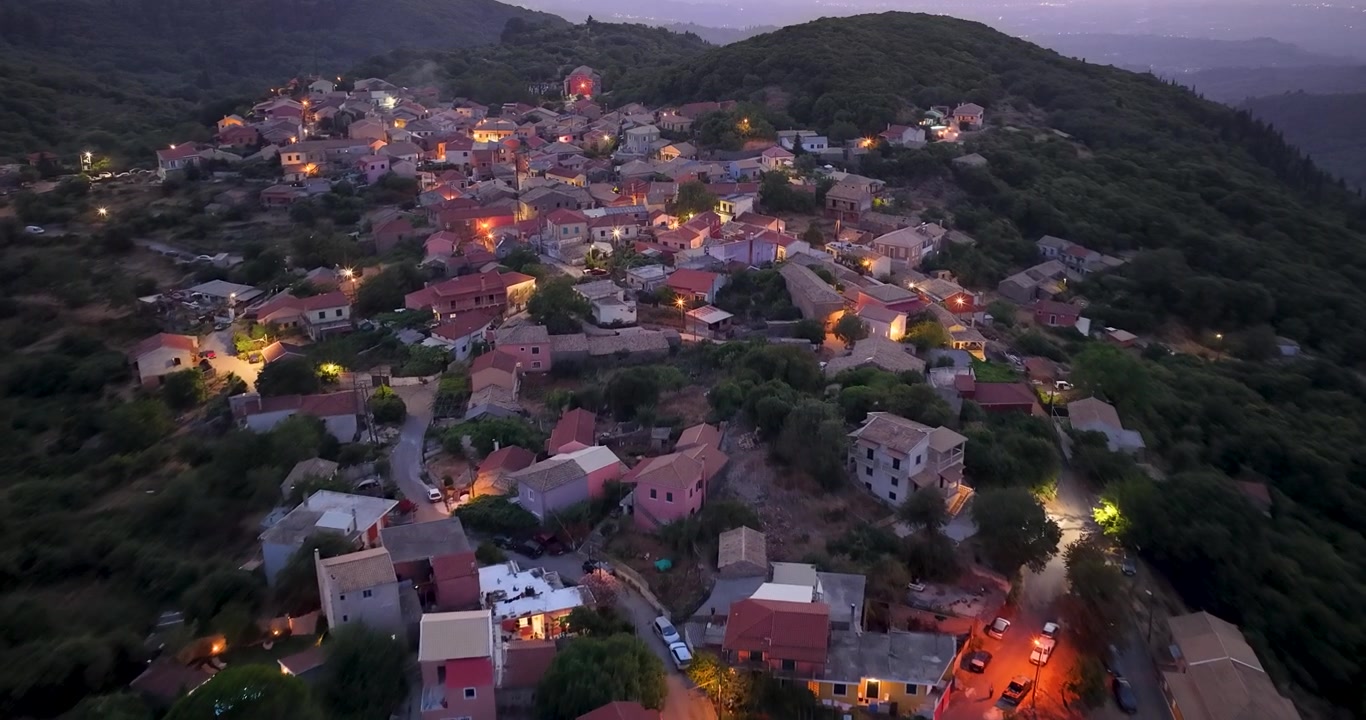 Ripresa con drone che sorvola le luci stradali del pittoresco villaggio di Sokraki al crepuscolo serale a Corfù Nord, Grecia. Movimento aereo sopra i tetti in tegole e il paesaggio montano durante la tranquilla ora del crepuscolo.