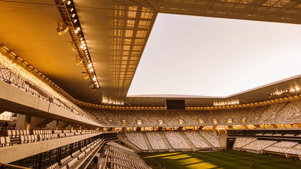 Pengambilan gambar dimulai, kamera miring memperlihatkan mangkuk stadion dan atap yang terbuka, menunjukkan pita bayangan di lapangan rumput
