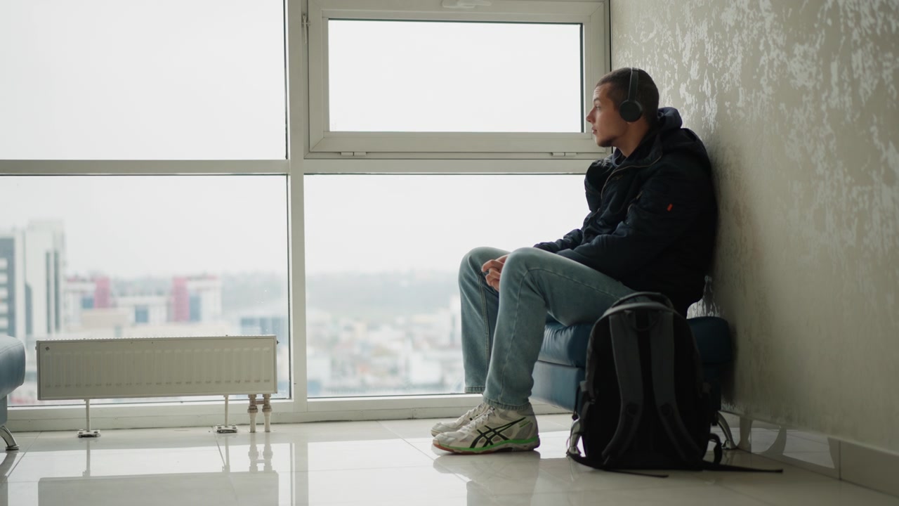 Joven sentado tranquilamente en una silla azul junto a grandes ventanales, con auriculares y ropa informal. El hombre parece estar esperando o descansando en un espacio interior con el paisaje urbano visible a través del cristal. Negro.